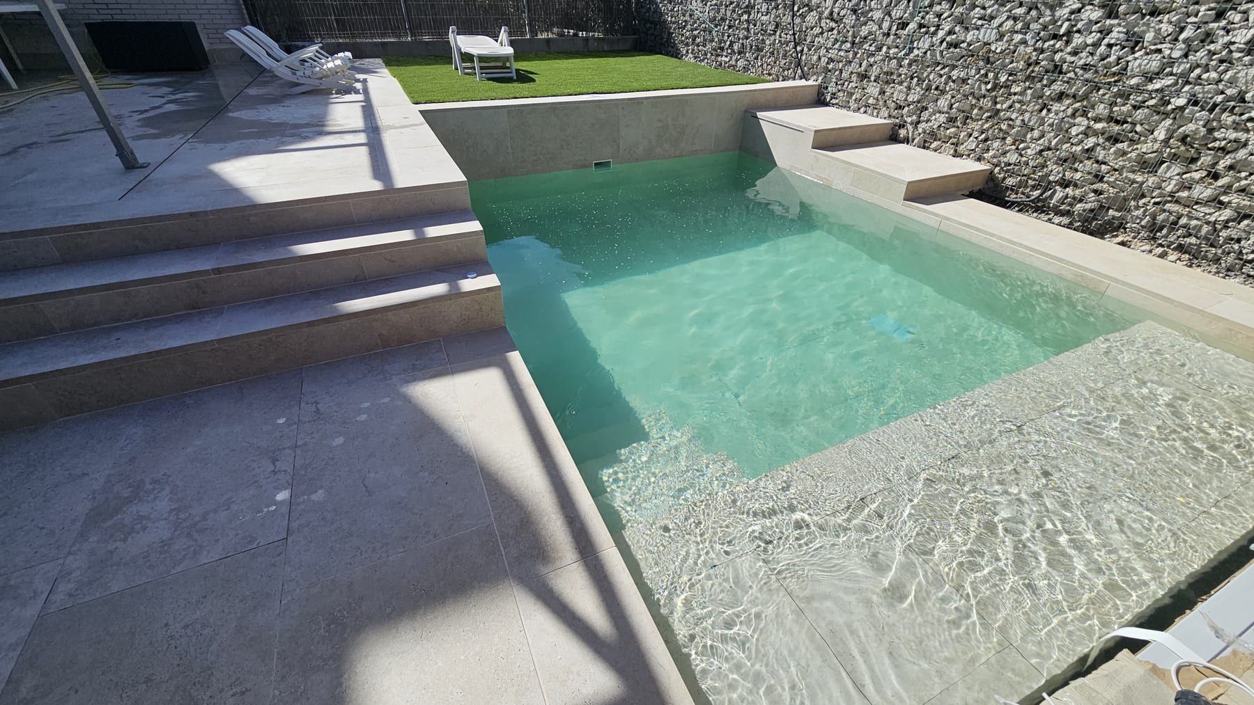 Piscina con escalera y playa junto a muro de piedra, con la escalera de ancho completo